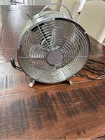 Front view of silver plug-in metal table fan showing protective grille and power cord.