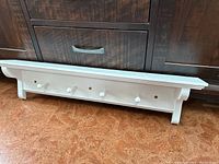 Front view of white wooden floating shelf with four hooks, placed on floor against dark furniture.