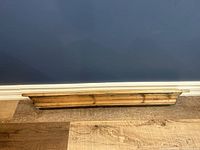 Front view of the antique wood floating shelf placed against the wall on the floor, showing the wood grain and carved decorative molding on the front edge.