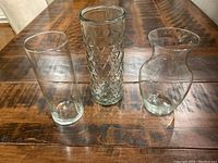 Photo showing three clear glass vases arranged on wooden table from above: one plain cylinder, one diamond pattern cylinder, one rounded with flared lip.
