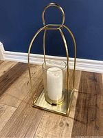 Gold metal framed lantern with white candle inside, standing on wood floor against blue wall, photo 1