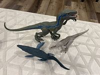 The three dinosaur figures on a beige surface showing size and color differences: Blue Velociraptor, grey Indominus Rex, and blue Mosasaurus.