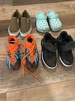 Four pairs of kids shoes laid on wooden floor: Vans slip-ons, blue Crocs, black Nike sneakers, and orange-blue soccer cleats.