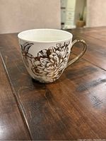 White ceramic mug with black floral design and gold handle, shown on dark wooden table