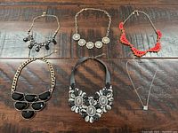 Top view of six necklaces laid out on wood surface showing black bead, rhinestone floral, red bead, black geometric, black and white floral statement, and clear pendant necklaces.