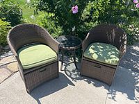 Two brown woven rattan armchairs with green cushions and small round glass-top rattan side table, outdoors on a concrete surface with surrounding greenery and pink flowers.