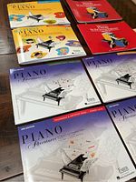 Set of seven children's piano lesson books: three with yellow and red covers featuring illustrations of children and a piano, and four with purple, white, and yellow covers including technique and performance books.