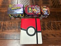 Photo showing three Pokémon card tins and the red and white Pokémon card binder closed on a wooden surface.