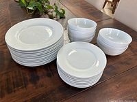 Stack of 11 white porcelain dinner plates, 10 white side plates, and 7 white bowls on a wooden table.
