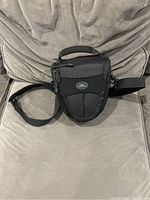 Front view of the black Roots camera bag with handle and shoulder strap, placed on a gray fabric surface.