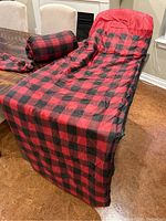 Full view of one red and black plaid sleeping bag set up on a chair with its matching compression sack beside it.