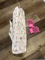 Full view of the white yoga mat carrier with yoga pose prints and pink yoga gloves beside it on floor.
