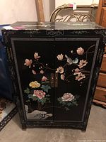 Front view of the cabinet showing the two black lacquered doors with painted floral and bird designs and brass handles.