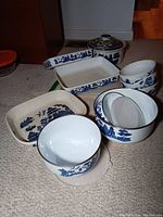 Photo showing multiple Blue Willow pattern dishes including two rectangular ceramic baking dishes, a lidded crock, and metal enamelware bowls and casserole dish grouped together.