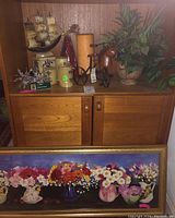 Photo of shelf with ship model, candles, Christmas star decorations, faux plant, and horsehead bicycle figure on top of wooden cabinet with framed floral print displayed in front.