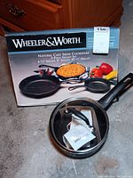 Photo of Wheeler & Worth cast iron cookware set box featuring three skillet images and a glass lid with handle in front of it, along with an attached tag.