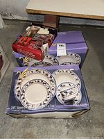 Two Lynns Brockton boxed stoneware dinnerware sets and one boxed Utica stainless steel flatware set shown in garage corner