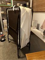 Folding rollaway cot folded upright, showing metal frame with black fabric bed surface and white mattress draped over it in a garage setting.