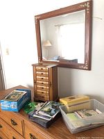 Wooden jewelry box with multiple front drawers and open side compartments on top of dresser with a large rectangular wooden floral painted mirror mounted on the wall above. Boxes of greeting cards and Saint Patrick's Day themed kerchief and beads are placed next to it.