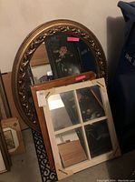 Round mirror with detailed gold-tone frame leaning against wall with two rectangular mirrors in front, one still in packaging with windowpane design