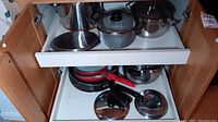 Full view of pots and pans arranged on two pull-out kitchen shelves revealing stainless steel and glass lids, multiple frying pans with red and black handles.