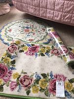 Photo showing two rectangular floral rugs and a rolled one on beige carpet, near a pink quilted bedspread.