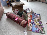 Photo showing rolled and flat floral and holiday themed door mats, coiled holiday lights, extension cords, and woven storage box with lid on carpeted floor