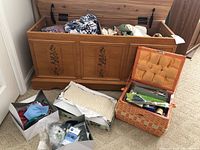Open wooden chest filled with assorted clothing, fabric pieces, and a wicker basket to the side containing sewing supplies. Two boxes on the floor with various fabric and notions.