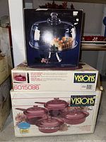 Three boxed sets stacked: Visions cranberry cookware with pots and lids, 4.0L covered oval roaster, and Tuscany pedestal cake stand & dome box on top.