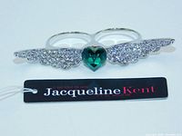 Double finger ring laid flat showing silver metal wings encrusted with clear crystals and a green heart-shaped crystal in the center, attached to two rings.