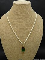 Front view of cream colored pearl necklace with rectangular green glass pendant coated in gold at the top suspended from a necklace bust.