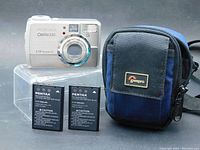 Front view of Pentax Optio 330 camera, two rechargeable batteries, and navy Lowepro camera case on clear plastic stand.