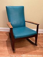 View of rocking chair showing teal faux leather seat and backrest with worn wood armrests and frame on hardwood floor.