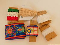 Photo showing all six cloth trimmings including braided ribbon, eyelet lace, zig zag trims, and embroidered ribbons arranged with length measurements noted on brown cardboard.