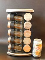Front angle photo showing 5 glass spice jars stacked vertically with metal lids embossed with spice names, attached to rotating rack with stainless steel base and top, soda can included for size reference.