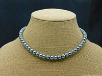 Full view of the necklace displayed on a beige textured bust, showing the smooth, lustrous single strand of silver/gray simulated pearls approximately 18 inches long.