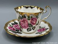 Front side view showing the scalloped edge teacup with pink rose pattern and thick gold trim on rim and handle, sitting on matching saucer with same pattern and gold rim.