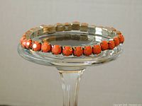 Coral-colored faceted stones set in a four-prong setting on a flexible, elastic metal expansion bracelet displayed on a clear glass stand.