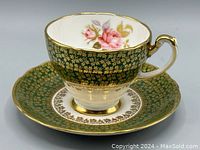 Side view of teacup on saucer showing dark forest green background, gold overlay pattern, pink rose decoration inside cup and on saucer center, gold rim and handle accents.