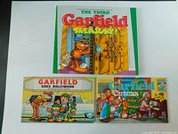 Three Garfield comic strip books positioned together showing their colorful front covers: The Third Garfield Treasury, Garfield Goes Hollywood, and Garfield Christmas.