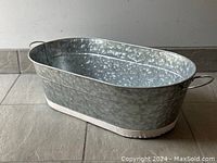 Large oblong galvanized metal tub on tiled floor showing side and inside interior, featuring textured finish and handles.