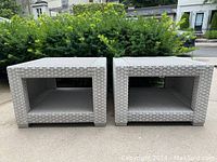 Pair of light gray resin outdoor side tables shown side by side facing front showing the rattan texture and open shelving underneath.