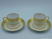 Two Grosvenor China demitasse cups and two matching saucers, white and pale yellow with gold gilt borders and intricate gold patterning on the edges.