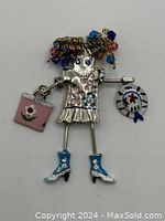 Front view of brooch showing silver-tone metal girl with multi-colored beaded curly hair, blue enamel boots, pink enamel purse, and white enamel hat.