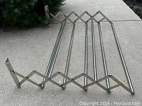 Wall-mounted stainless steel expandable clothes drying rack shown fully expanded on concrete surface with greenery background.