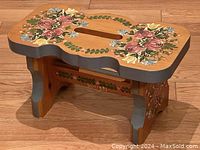 Footstool showing overall handpainted floral design on top surface, including symmetrical rose clusters, leaves, and small flowers, with a handle cutout in the middle and scalloped edges.