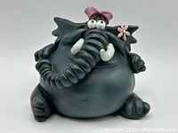 Front view of dark green ceramic elephant piggy bank featuring exaggerated rounded body and curled trunk with white tusks, pink bow on head, googly eyes, and pink flower detail.