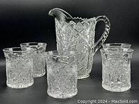 Set of one pressed glass jug and five matching glasses with clear intricate floral and diamond patterns against a black background.