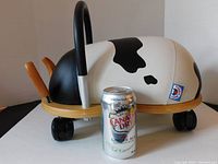 Side profile view showing black and white cow pattern body, wooden base with four black wheels, and Canadian Dry Ginger Ale can for size reference.