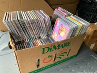 Photo showing a cardboard box packed with numerous used CDs standing upright and stacked, housed in a DiMare brand tomato box.